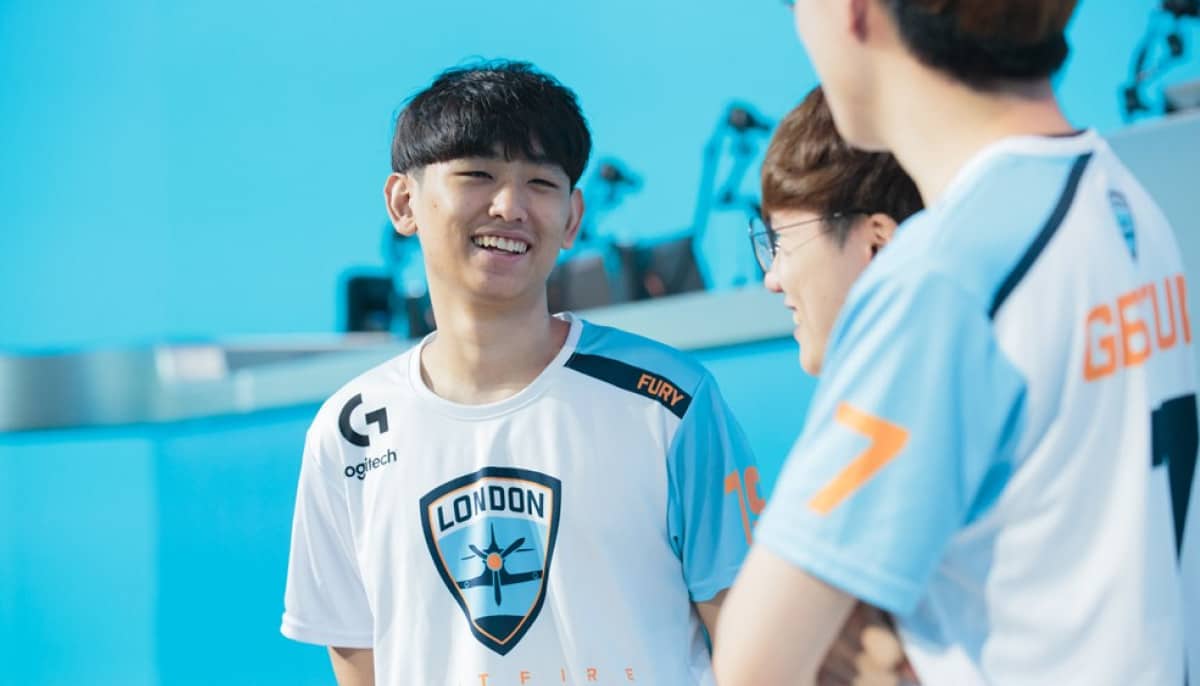 Three esports players wearing light blue and white jerseys stand together, smiling and chatting